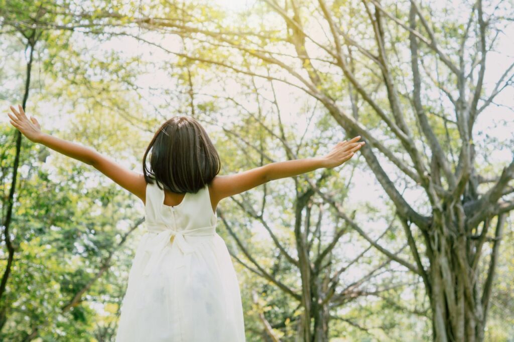A young girl in a white dress enjoys the outdoors with arms outstretched under the sunlit forest canopy.